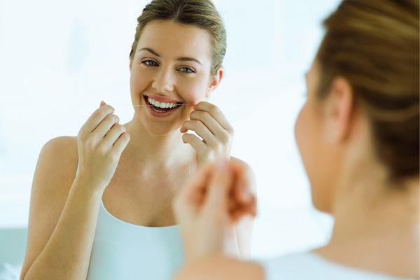 La mejor forma de prevenir el mal aliento Mujer sonriendo frente al espejo mientras usa hilo dental.